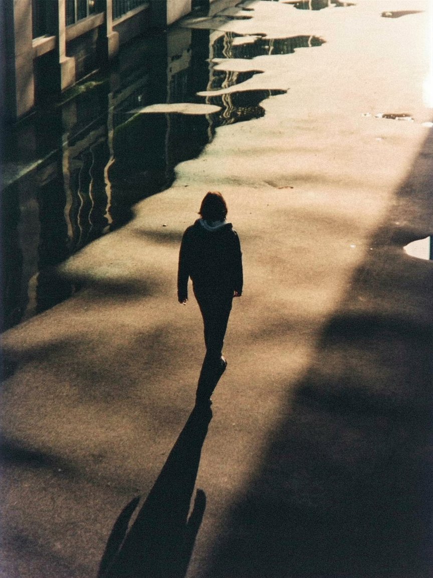 একটি বাঁকা রাস্তার দৃশ্য যেখানে এক একা মানুষ পিছনের দিকে হাঁটছে, তার ছায়া অসম্ভব লম্বা, মাটি পানির মতো নড়ছে, দৃষ্টিভঙ্গি বিকৃত, ভূমিকম্পের অস্থিরতা এবং একাকীত্বের প্রতীক﻿