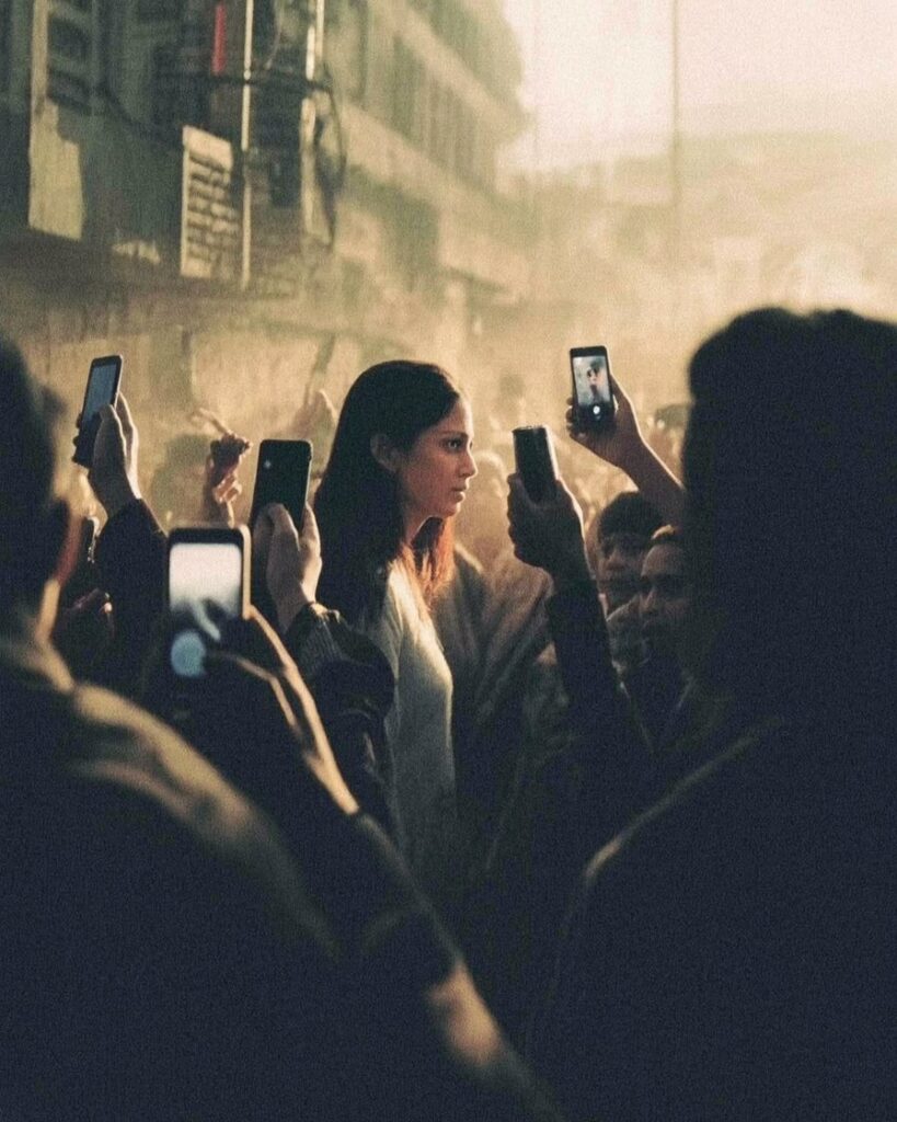 ঢাকা শহরের এক ভিড়ভাট্টা রাস্তায় মানুষজন এক নারীর প্রতি বিচার ছুঁড়ে দিচ্ছে — কেউ ভিডিও করছে, কেউ তাকিয়ে আছে। কারও মুখ স্পষ্ট নয়, সবার চোখে একই অন্ধকার। ছবিটি প্রতীক — এক সমাজের, যেখানে সবাই মুখোশ পরে বেঁচে থাকে, আর কেউই সম্পূর্ণ নিষ্পাপ নয়।