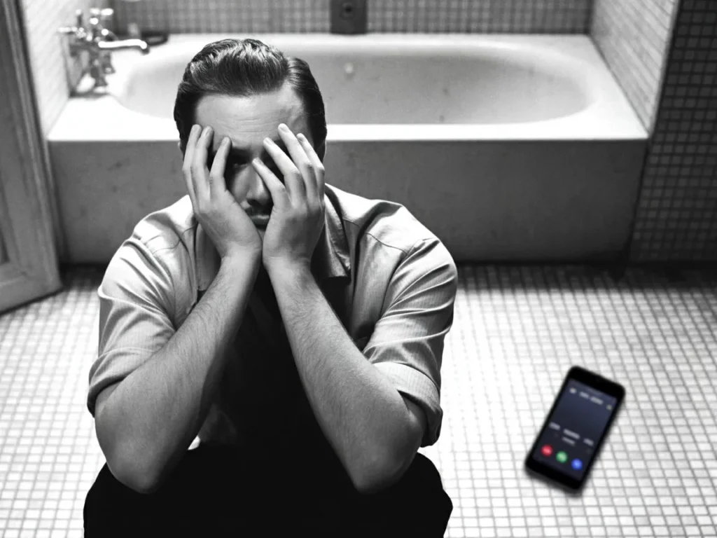 A photorealistic, cinematic shot of a man in a rumpled shirt sitting on a bathroom floor at 3 am, back against the bathtub and knees to chest, one hand covering his face in grief, with a glowing phone screen showing an unanswered call on the tiles under harsh overhead lighting.